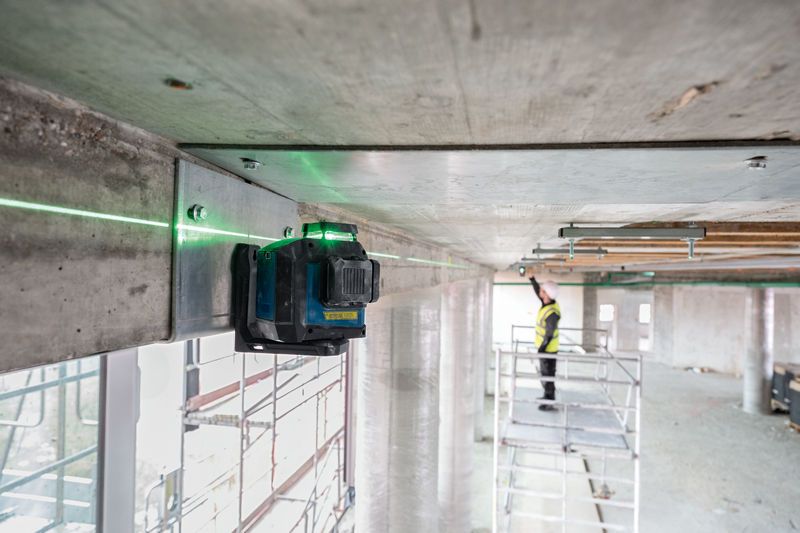 Herramienta niveladora láser proyecta líneas verdes en un techo, persona con equipo de seguridad ajusta la alineación.