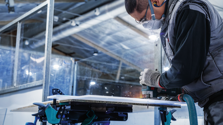 Una persona que usa equipo de seguridad utiliza una herramienta eléctrica para cortar un panel de madera.
