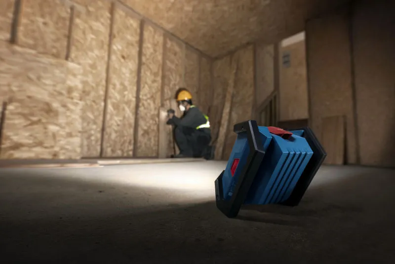 Una persona que usa equipo de seguridad instala paneles de yeso, iluminada por una luz de trabajo inalámbrica.