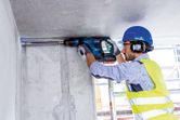Una persona que usa equipo de seguridad perfora un techo de hormigón utilizando un martillo perforador inalámbrico.