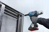 Una persona que usa equipo de seguridad fija un panel de yeso a un marco de metal con una herramienta eléctrica.