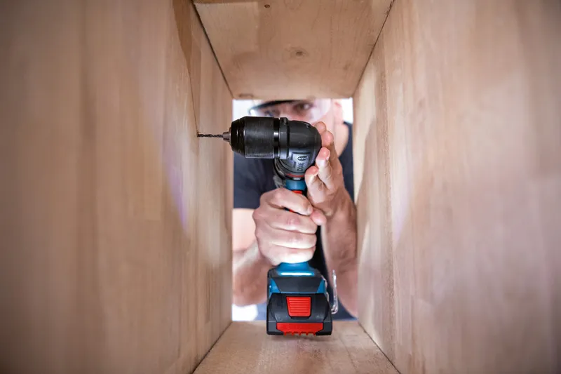 Una persona que usa equipo de seguridad perfora un panel de madera dentro de un gabinete.