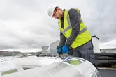 Una persona que usa equipo de seguridad usa un taladro eléctrico en un conducto metálico grande al aire libre.
