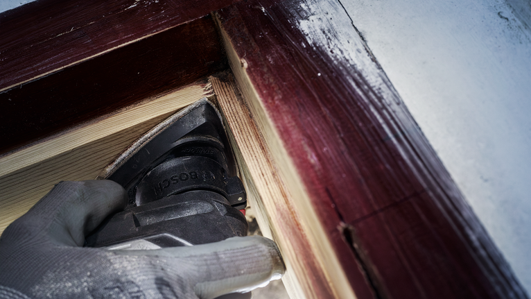 Una persona que usa equipo de seguridad lija madera cerca del marco de una ventana pintado.