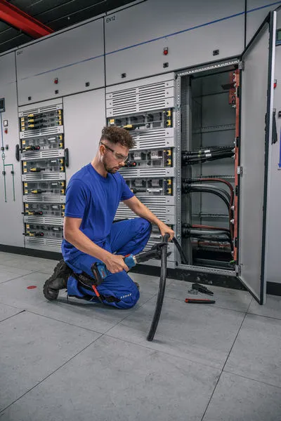 Una persona que usa equipo de seguridad corta un cable grueso frente a un panel eléctrico abierto.