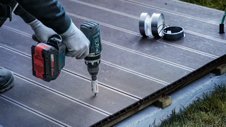 Una persona que usa equipo de seguridad utiliza un taladro inalámbrico para hacer un agujero en la terraza.