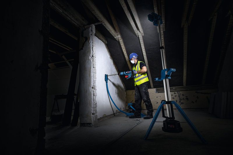 Una persona equipada con equipo de seguridad taladra una pared, iluminada por una luz de obra inalámbrica.
