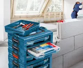 Naskládané bedny s nářadím s otevřenou zásuvkou s různými spojovacími prvky, v pozadí osoba v bezpečnostním vybavení.