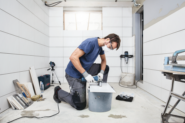 Osoba v bezpečnostním vybavení vyvrtá díru do dlaždice uvnitř obložené místnosti.