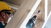 Osoba v bezpečnostním vybavení vyvrtá díru do dřevěných trámů.