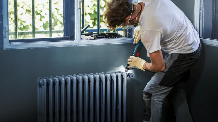 Osoba v bezpečnostním vybavení používá akumulátorovou vrtačku k instalaci radiátoru.