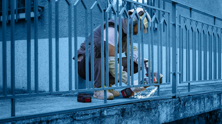 Osoba v bezpečnostním vybavení používá elektrické nářadí k řezání kovového plotu.