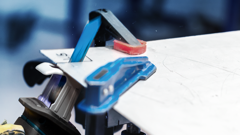 Osoba v bezpečnostním vybavení řeže plechový materiál elektrickým nářadím na pracovním stole.