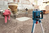 Laserový nivelační přístroj na stativu vedle pracovníků v bezpečnostním vybavení, kteří lijí beton.