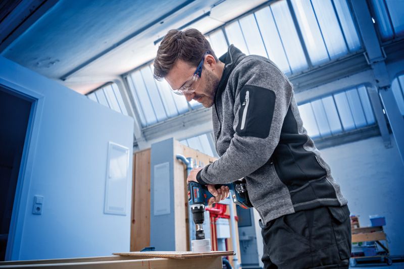 Osoba v bezpečnostním vybavení používá akumulátorovou vrtačku k vyvrtání otvoru do dřeva.