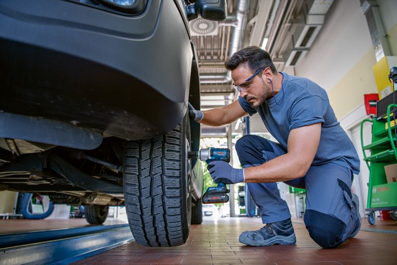 Osoba v bezpečnostním vybavení utahuje matice na kole automobilu pomocí akumulátorového rázového utahováku.