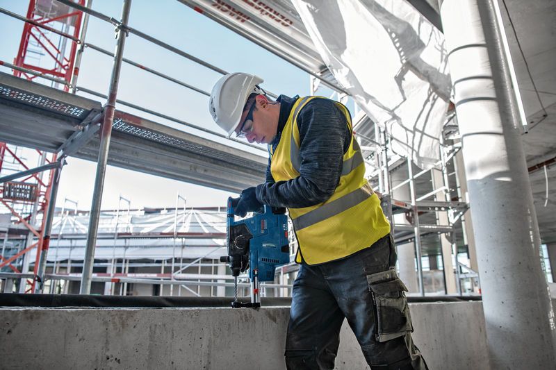 Osoba v bezpečnostním vybavení vrtá do betonu na staveništi.