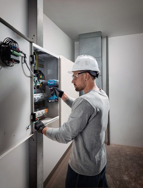 Osoba v bezpečnostním vybavení používá akumulátorovou vrtačku na elektrickém panelu.