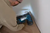 Osoba v bezpečnostním vybavení používá akumulátorovou hřebíkovačku na dřevo k upevnění lišt k podlaze.