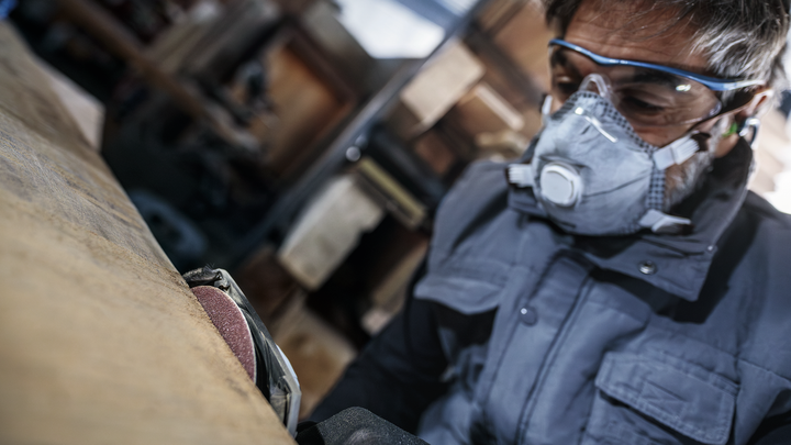 Osoba v bezpečnostním vybavení brousí dřevěný povrch elektrickým nářadím.