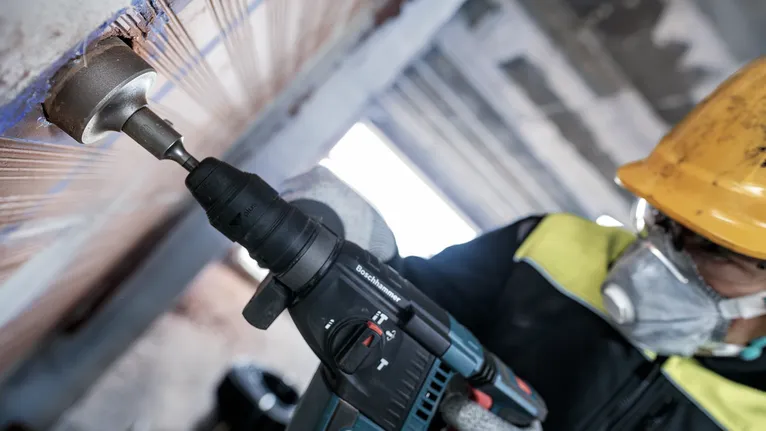 Osoba v bezpečnostním vybavení vrtá do zdi elektrickým nářadím.