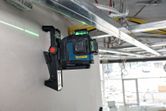 Laserový nivelační přístroj namontovaný na zdi, promítající zelený laserový paprsek napříč staveništěm.