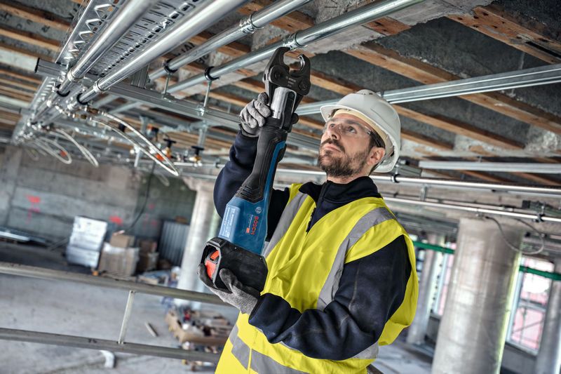 Osoba v bezpečnostním vybavení používá akumulátorový lis na nadzemních trubkách.