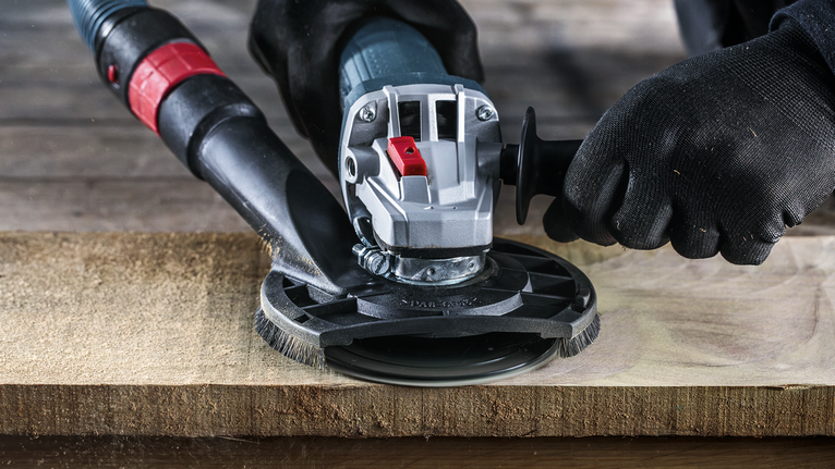 Osoba v bezpečnostním vybavení brousí dřevěný povrch elektrickým nářadím.