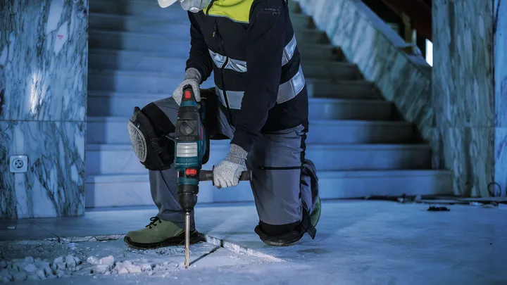 Osoba v bezpečnostním vybavení vrtá do betonové podlahy uvnitř.