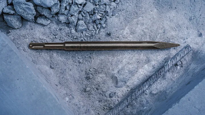Špičatý dláto spočívající na zaprášeném betonu poblíž odkryté výztuže.