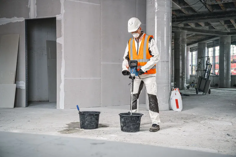 Osoba v bezpečnostním vybavení používá míchací mechanismus k míchání materiálu v kbelíku.