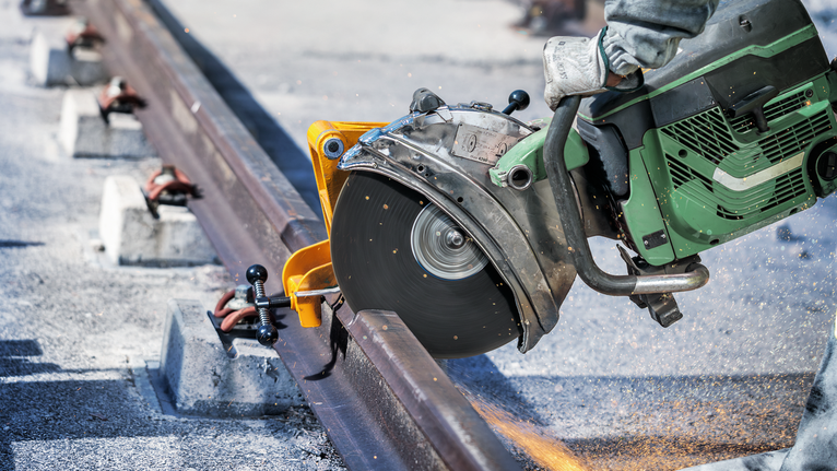 Řezací pila řeže ocelovou kolejnici, pracovník v bezpečnostním vybavení.
