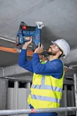 Osoba v bezpečnostním vybavení používá akumulátorovou hřebíkovačku na beton na stropě.