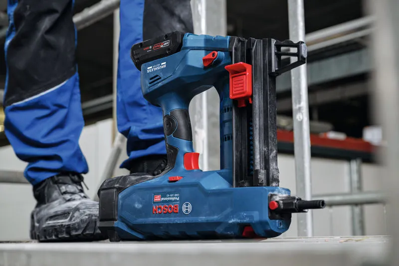 Akumulátorová hřebíkovačka na beton odpočívající na stavební ploše poblíž osoby v bezpečnostním vybavení.
