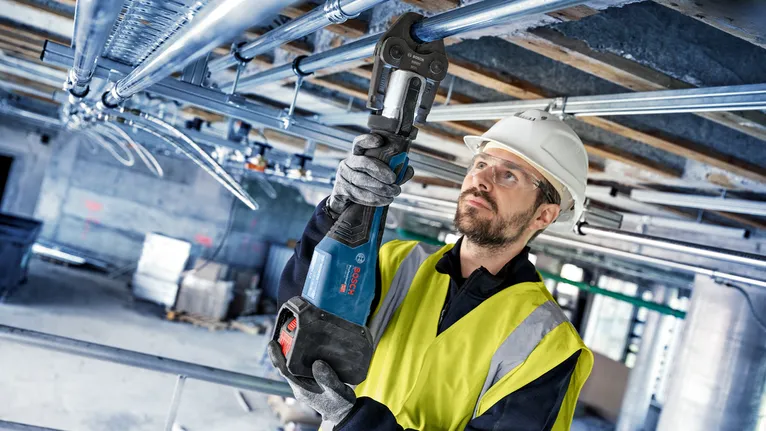 Osoba s bezpečnostním vybavením používá elektrické nářadí k zajištění kovových trubek nad hlavou.