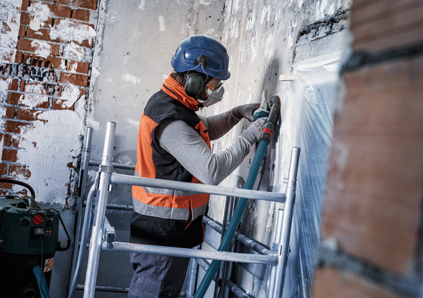 Osoba v bezpečnostním vybavení brousí betonovou zeď na lešení.