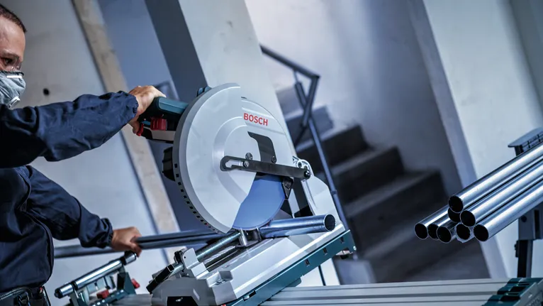 Osoba v bezpečnostním vybavení řeže kovové trubky kotoučovou pilou.