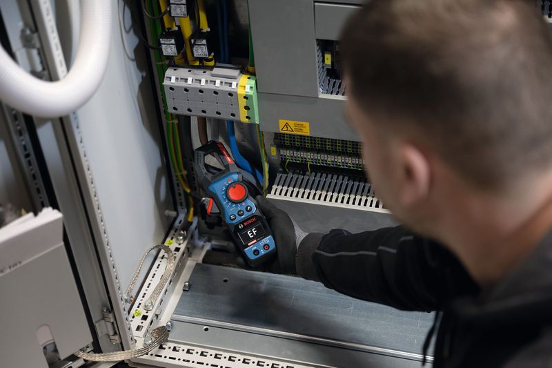 Osoba v bezpečnostním vybavení používá klešťový měřič ke kontrole elektrického rozvaděče.