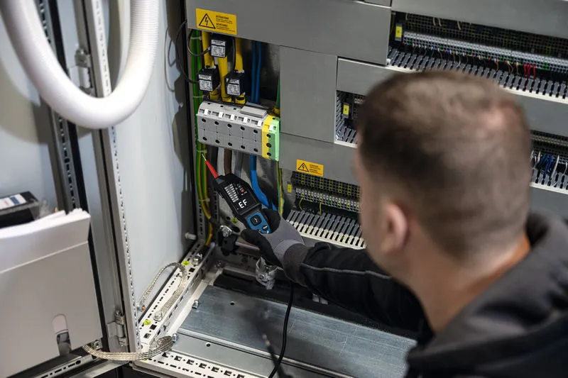 Osoba v bezpečnostním vybavení používá zkoušečku napětí na elektrickém rozvaděči.