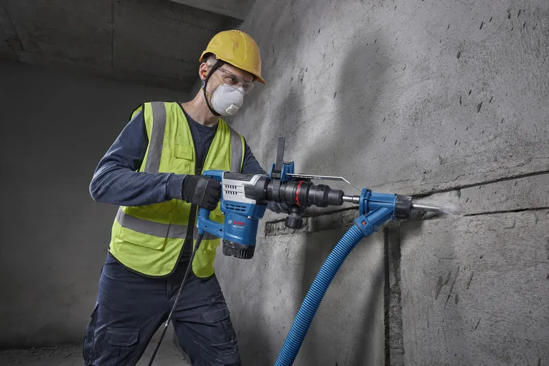 Osoba v bezpečnostním vybavení vrtá do betonové zdi pomocí elektrického nářadí.