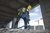 Osoba v bezpečnostním vybavení bourá beton bouracím kladivem.