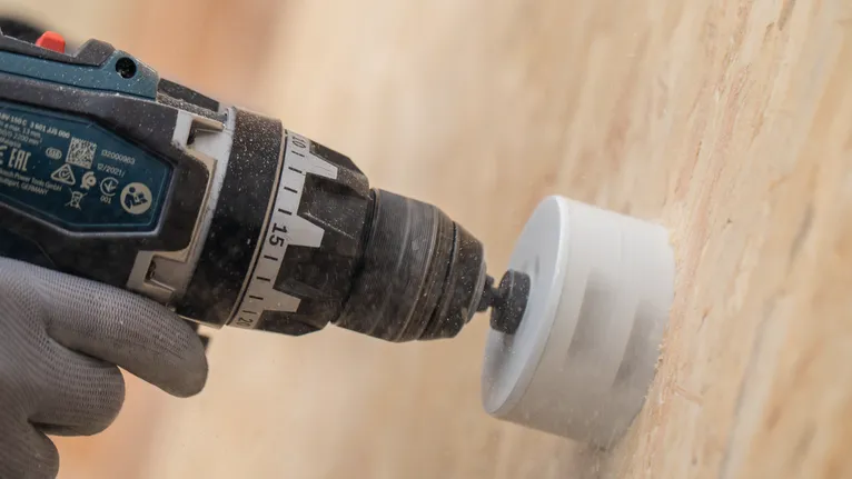 Osoba v bezpečnostním vybavení vyvrtá kruhový otvor do dřevěné zdi.