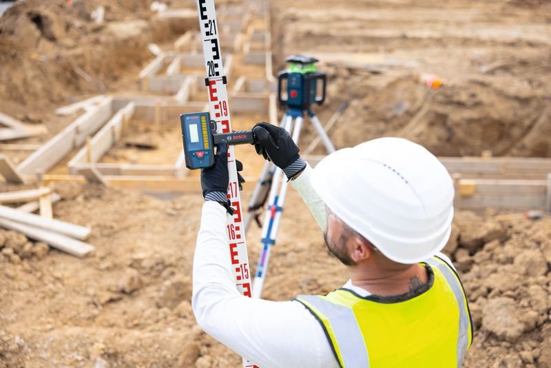 Osoba v bezpečnostním vybavení používá na staveništi laserový přijímač na nivelační lati.