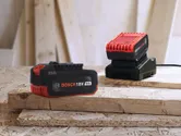 Dobíjecí baterie a nabíječka do elektrického nářadí ležící na naskládaných dřevěných deskách.