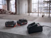 Dvě dobíjecí baterie do nářadí a nabíječka na betonovém povrchu ve staveništi.