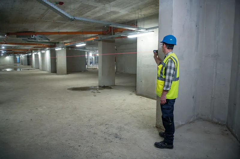 Osoba v bezpečnostním vybavení používá laserový nivelační přístroj v betonové budově.