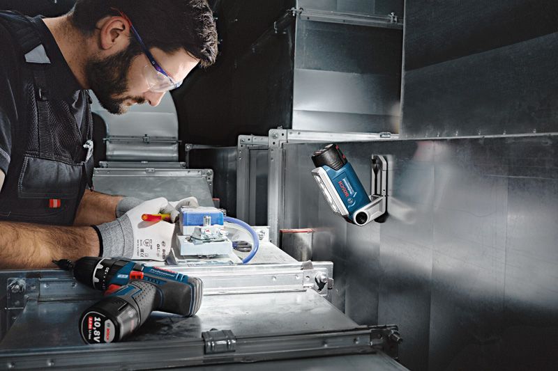 Eine Person mit Sicherheitsausrüstung arbeitet mit elektrischen Leitungen unter einer kabellosen Arbeitsleuchte.