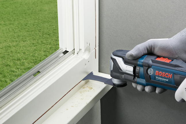 Eine Person, die Schutzausrüstung trägt, bearbeitet einen Fensterrahmen mit einem Multifunktionswerkzeug.