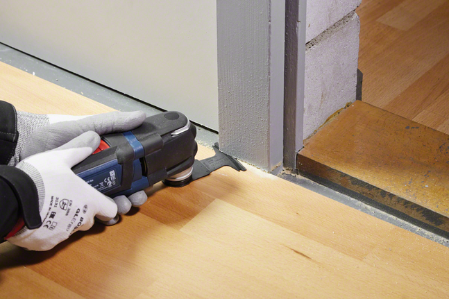 Eine Person, die Schutzausrüstung trägt, schneidet mit einem Elektrowerkzeug Laminatboden in der Nähe eines Türrahmens.