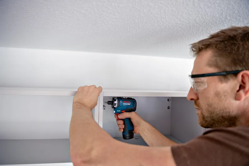 Eine Person mit Schutzausrüstung baut mit einem Akkuschrauber einen Schrank zusammen.